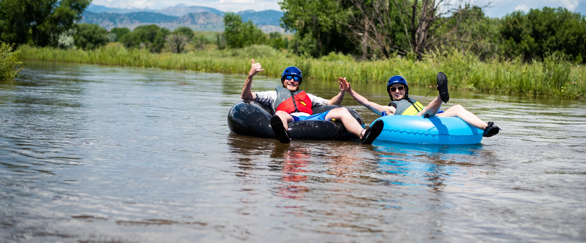 Two smiling people float a river on inflatable tubes.