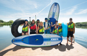 Smiling group holds inflatable paddleboards and tubes on a dock.