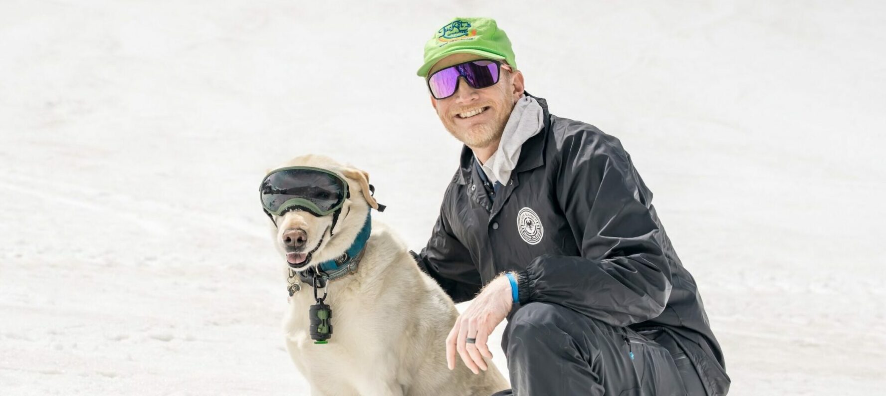 Man kneeling on snow next to a dog with goggles smiles at the camera.